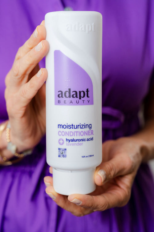 Person holding a bottle of Adapt Beauty moisturizing conditioner against a purple background